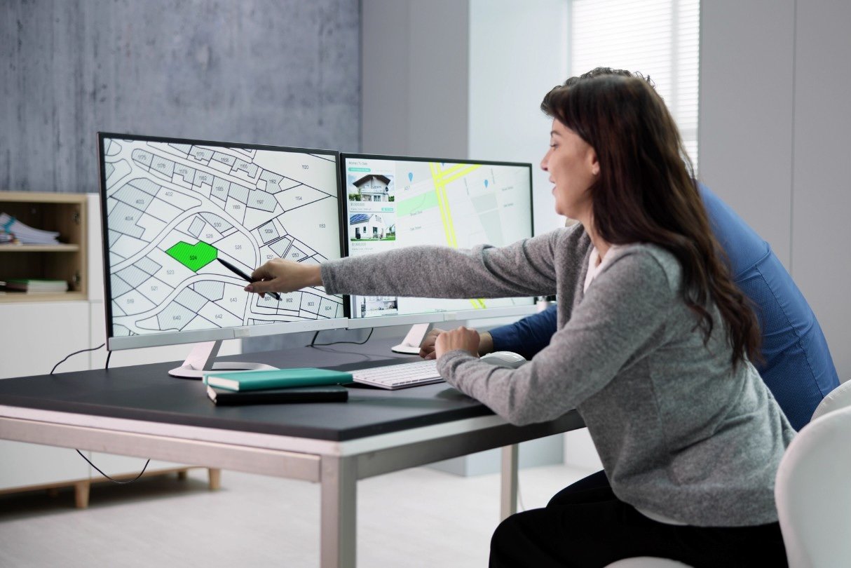 Two women sitting at a desk with dual monitors displaying maps and property listings, one using a pointer to highlight a specific lot on the map.