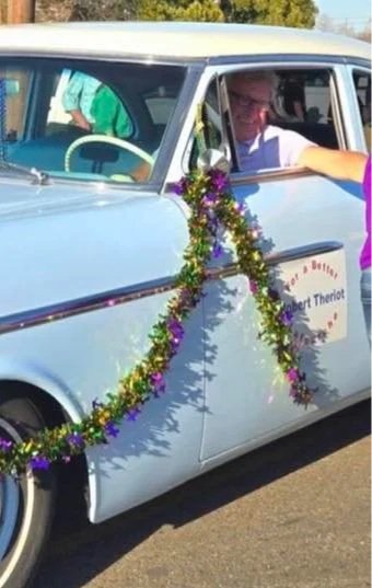 A vintage light blue car decorated with purple and gold tinsel and flowers, parked outdoors. Two people are inside, one smiling and another leaning out of the window, during what appears to be a parade or festive event.