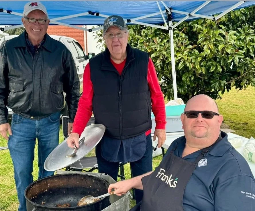 Three men at an outdoor barbecue under a blue canopy, one man grilling food, two others standing nearby, all smiling