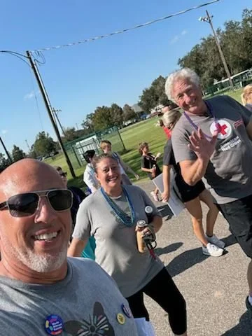 Group of people walking outdoors on a sunny day, some smiling and waving, near a sports field surrounded by trees and power lines.