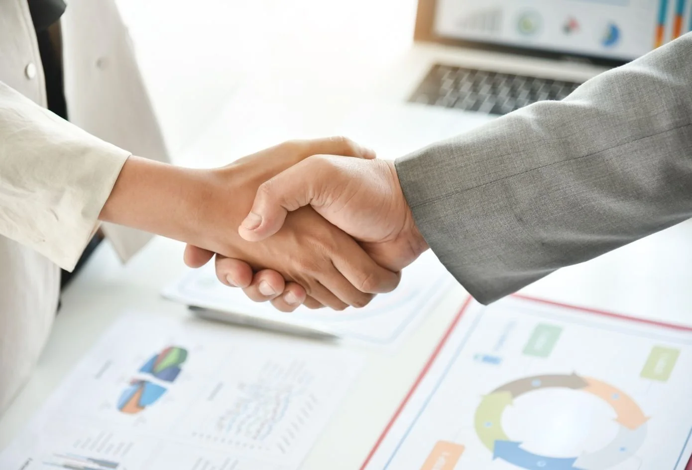 Two people shaking hands over a desk with documents and a laptop.