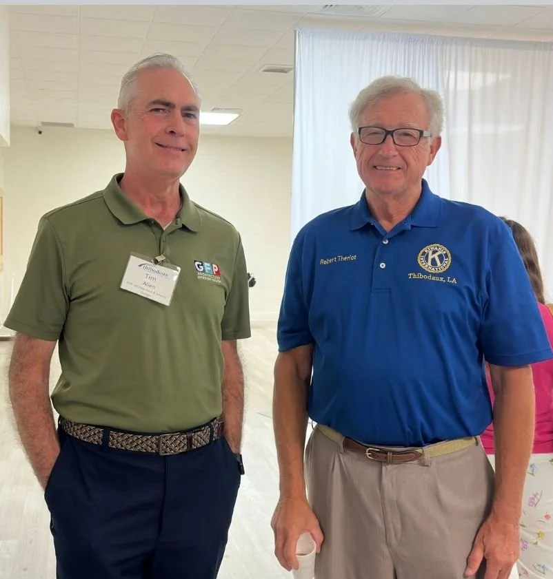 Two older men standing indoors, smiling at the camera. The man on the left wears a green polo shirt with name tag and emblem, navy pants, and a patterned belt. The man on the right wears a blue polo shirt with embroidered logo and text, beige pants, and glasses, holding a white cup in his right hand.