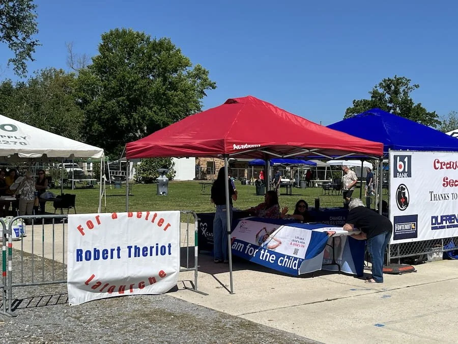 Local event with tents and tables, one table has a banner for a charity called 'Lymphomaniacs for the child.' There are people sitting and talking under the tents, with a clear blue sky and trees in the background.