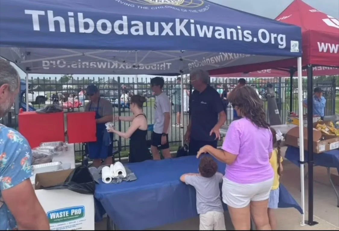People gather at a food stand with a blue canopy reading 'ThibodauxKiwani.org' and a red canopy. The stand is serving food, and customers are waiting in line and being served.