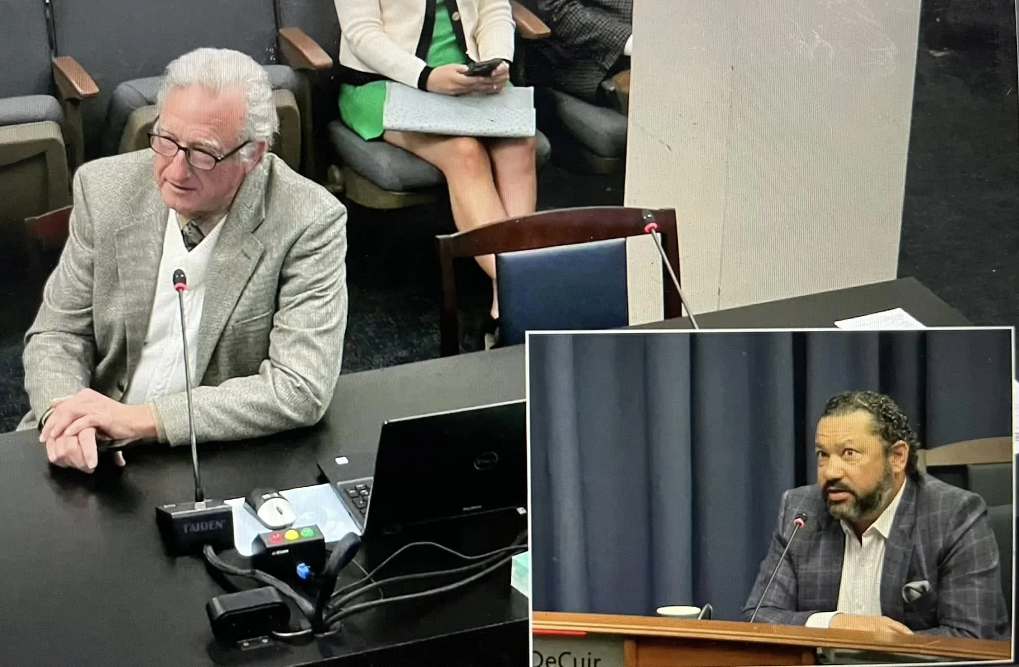 Two men sitting at a conference table during a formal meeting. They are involved in discussions with microphones and laptops in front of them. Behind them, a woman is seated with a notepad on her lap, using her phone. The setting appears to be a prof