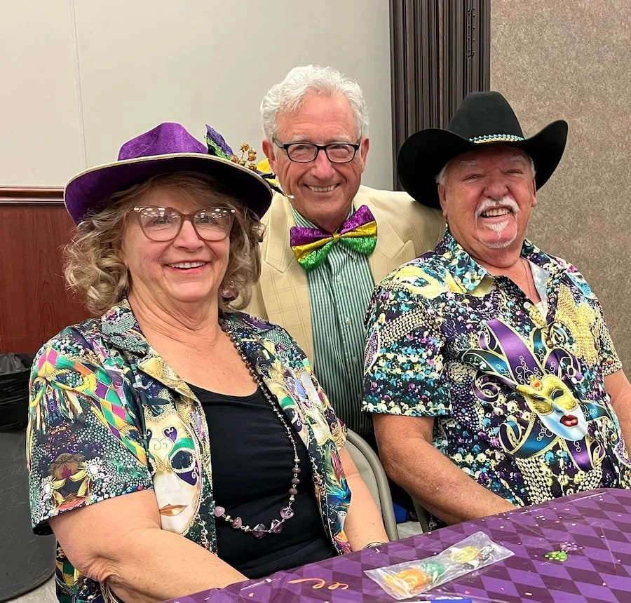 Three people dressed festively for Mardi Gras, wearing colorful masks and hats, sitting at a table with a purple tablecloth.