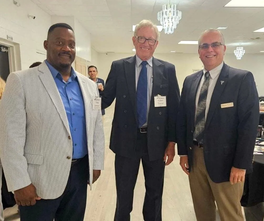Robert Theriot stands smiling between two men at a community event, all dressed in business attire with name tags visible in a bright meeting room with chandeliers overhead.