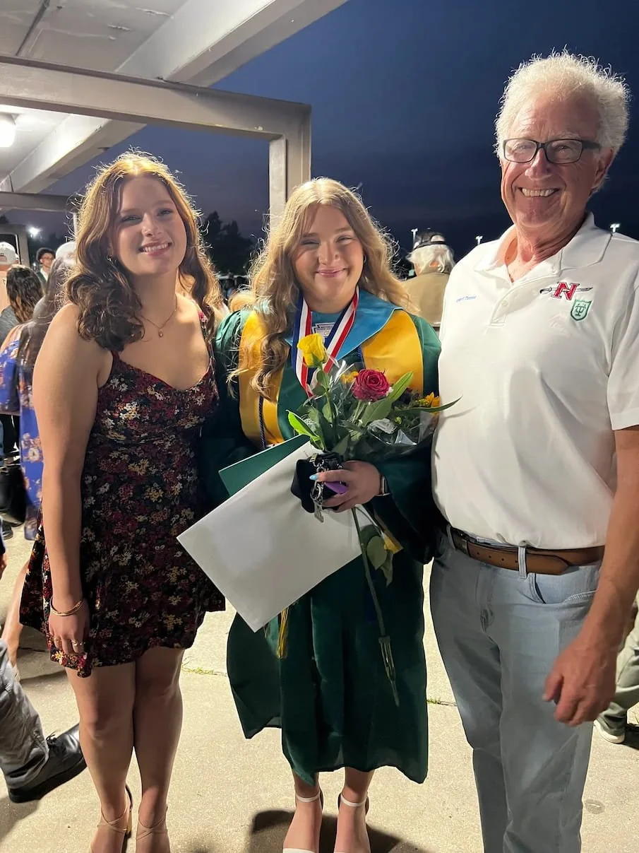 Robert Theriot stands smiling beside his granddaughters at a graduation celebration, one wearing a green cap and gown with medals and holding flowers and a diploma while attendees gather in the background at dusk.