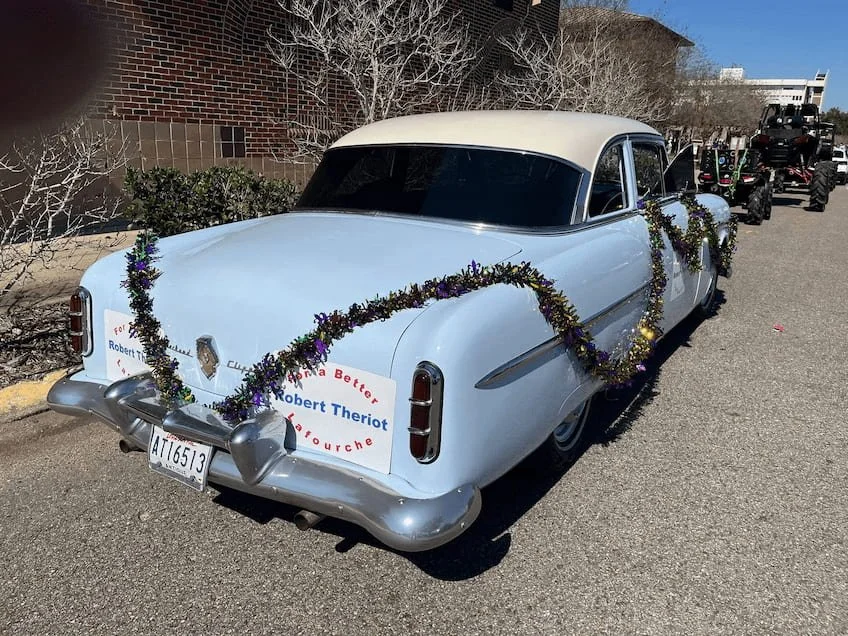 A vintage white car decorated with purple and gold garlands parked on a street during a parade or celebration.