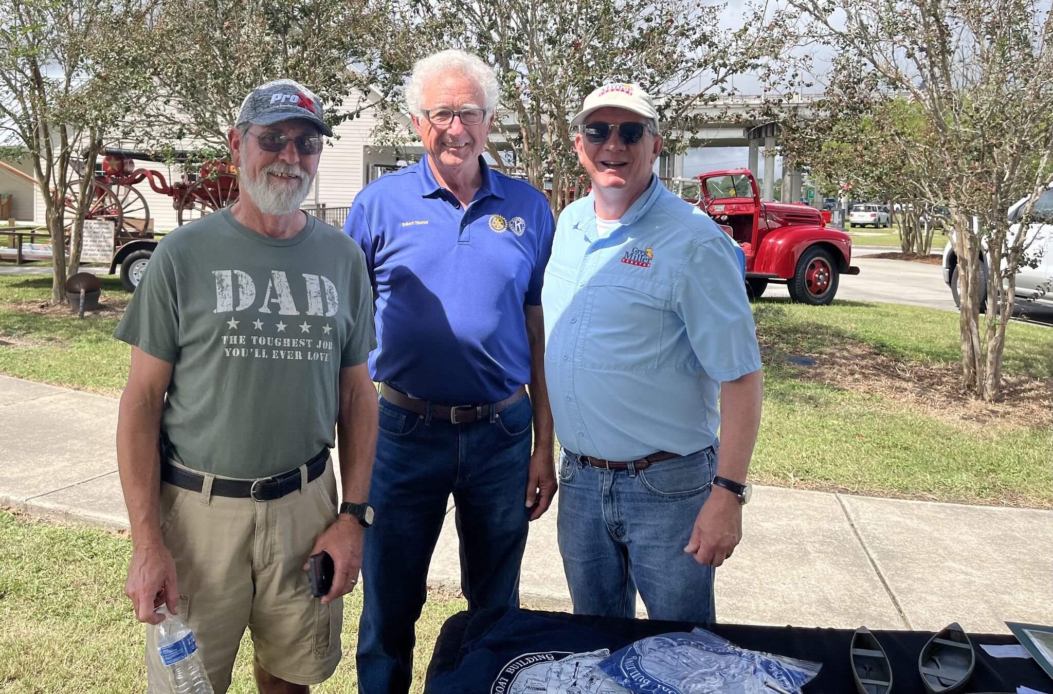 Three men standing outdoors in front of trees and vintage red trucks, smiling at the camera. The man on the left has a beard and glasses, wearing a gray T-shirt and a cap. The man in the middle has white hair, glasses, and wears a blue polo shirt with a Rotary emblem. The man on the right is wearing sunglasses, a white cap, and a light blue shirt.