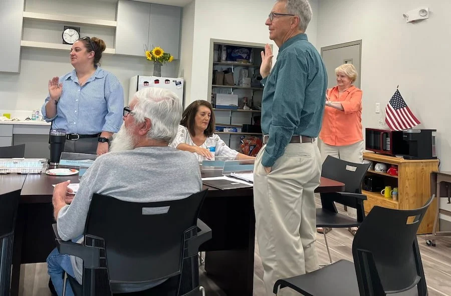 Group of older and younger adults in a casual office meeting room with American flag, flowers, and kitchen in background. Some people are standing for a pledge or oath including Robert Theriot.