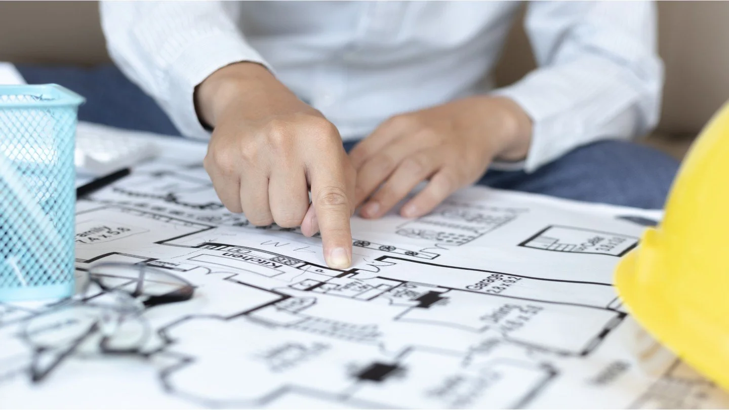 Person pointing at a floor plan with a finger, with a yellow construction helmet nearby.