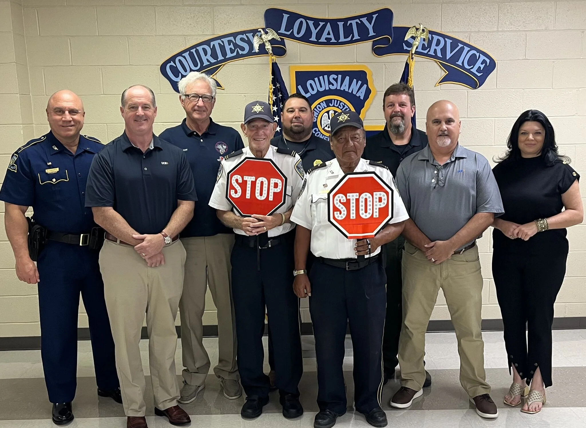 A group of nine people, mostly men, standing in front of a Louisiana police or sheriff's office emblem and banner that reads 'Courtesy Loyalty Service.' Four of them, two men, are holding stop signs.