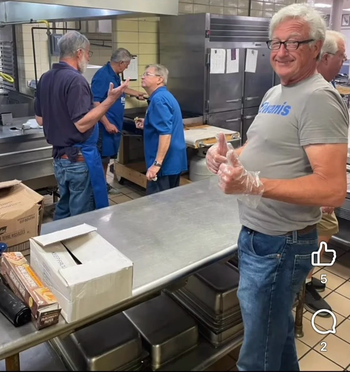 Group of people working in a commercial kitchen, some wearing blue aprons, one person in the foreground is smiling and giving a thumbs up, wearing gloves and a gray T-shirt, with boxes and kitchen equipment on metal tables.