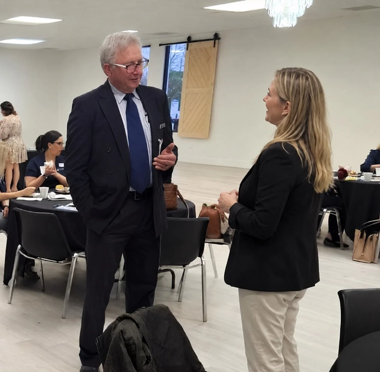 Robert Theriot and a woman engaged in conversation during a professional event in a bright, modern conference room. The man is wearing a dark suit with a blue tie, and the woman is dressed in a black blazer and light-colored pants.
