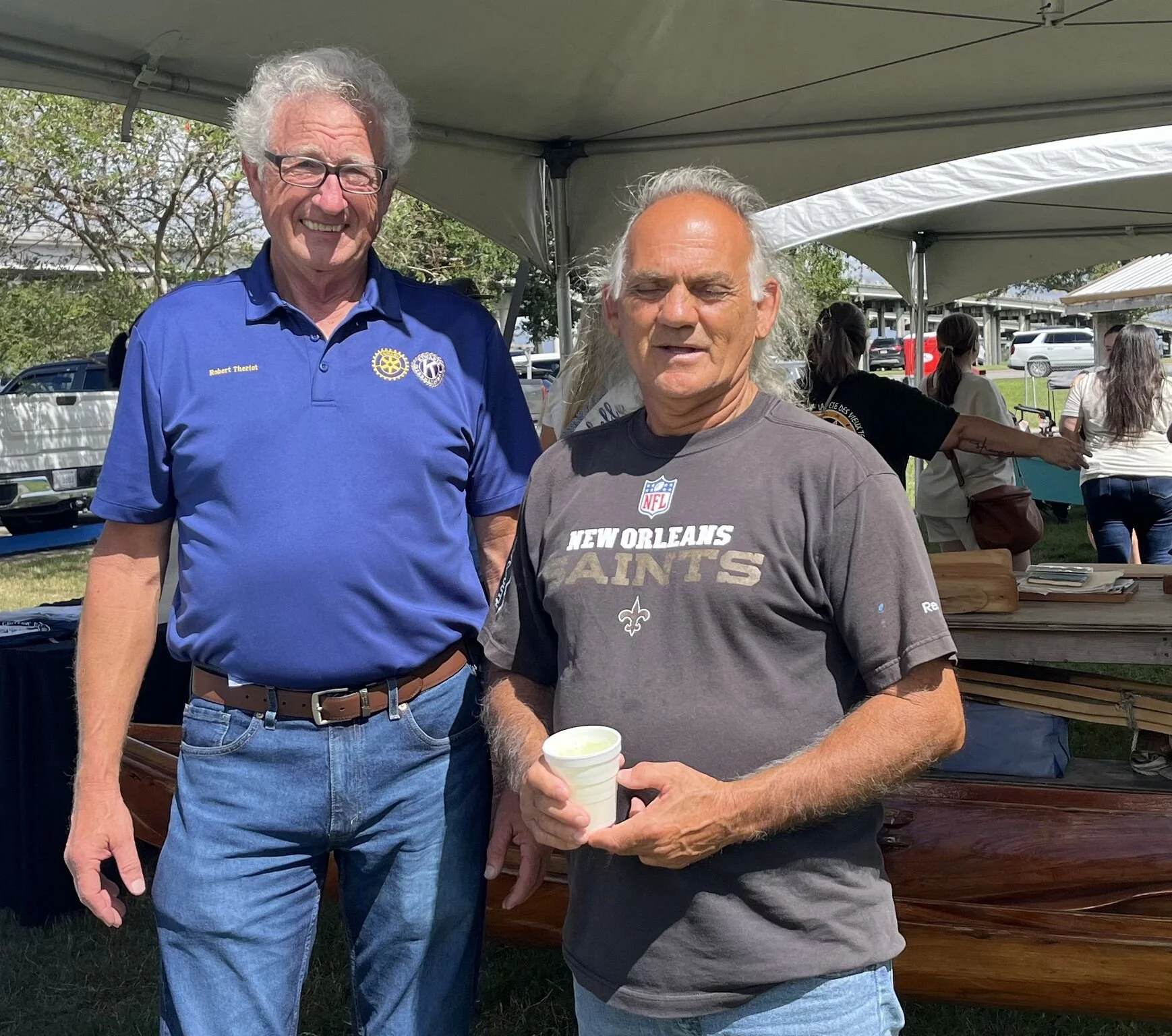Two older men standing under a tent at an outdoor event. The man on the left is wearing glasses, a blue polo shirt with a Rotary International logo and text, jeans, and a brown belt, smiling at the camera. The man on the right has long gray hair, is wearing a gray NFL New Orleans Saints T-shirt, holding a cup in his right hand, and appears to be mid-conversation. In the background, there are other people, some tables, and trees.