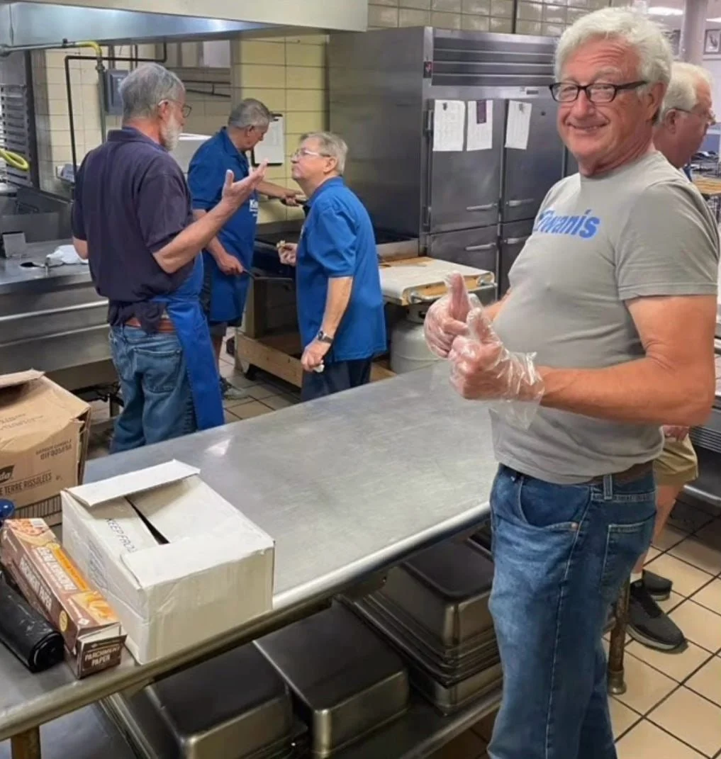 Group of people in a kitchen, some wearing blue aprons, chatting and working, Robert Theriot in the foreground volunteering, giving a thumbs-up and wearing gloves.