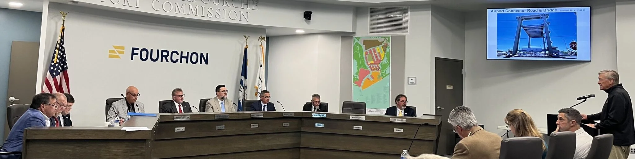 People seated at a long desk in a meeting room, with a speaker standing at a microphone on the right, and a large display screen on the wall showing an image of the Airport Connector Road & Bridge. The room has flags, a large Fourchon sign, and a map