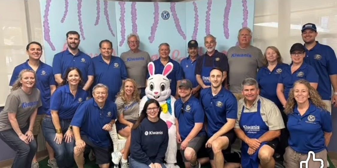 Group photo of people, some wearing Kiwanis shirts, with a person in an Easter Bunny costume, in front of a pastel-colored backdrop with hanging decorations.