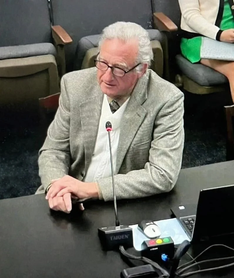 Robert Theriot wearing a gray blazer and a white sweater, sitting at a table speaking into a microphone. He has his hands clasped on the table and appears to be in a formal setting or meeting.