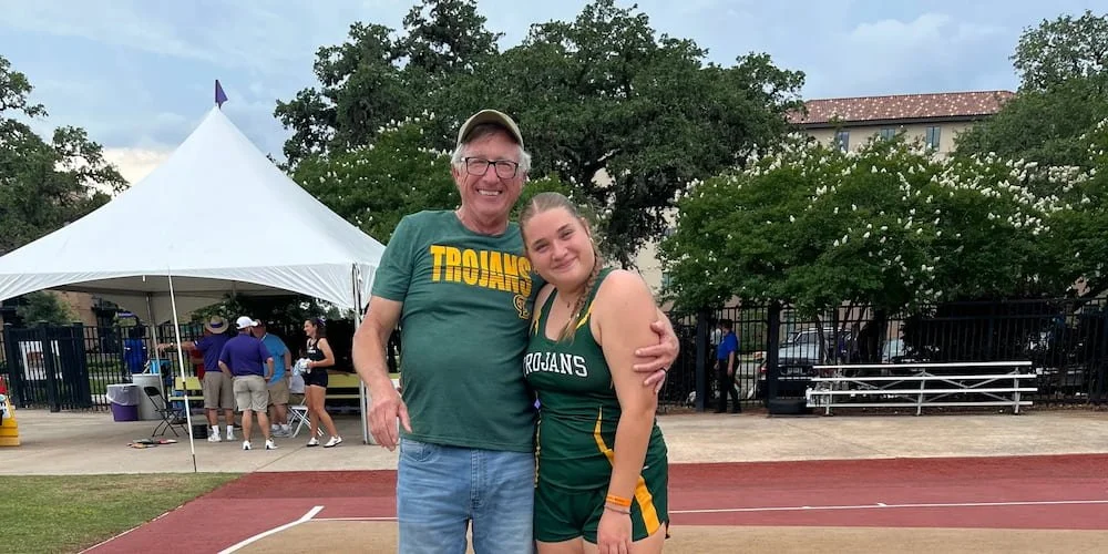 Robert Theriot and his granddaughter hugging on a running track at an outdoor event with a white tent and trees in the background.