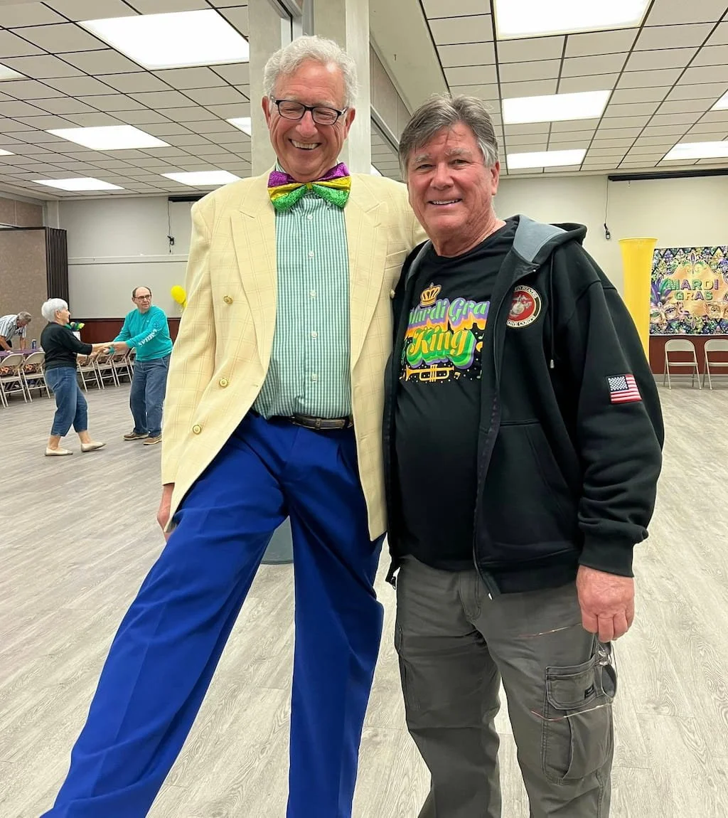 Two men standing together, smiling at the camera. The man on the left wears a yellow plaid blazer, green shirt, colorful bow tie, and blue pants. The man on the right wears a black hoodie with a Mardi Gras theme and gray cargo pants. In the backgroun
