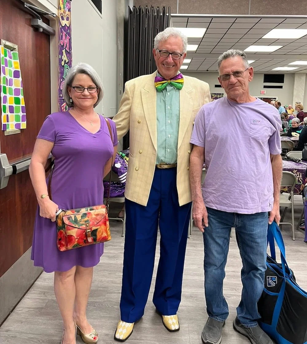 Three people standing together in an indoor event. The woman on the left is wearing a purple dress, glasses, and holding a floral handbag. The man in the middle is wearing a yellow plaid blazer, a colorful bow tie, and patterned shoes. The man on the