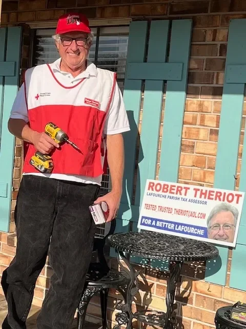 A man dressed in a Red Cross shirt and red cap standing outside a house with a brick wall and teal window shutters, holding a cordless drill and a measuring tape. There is a campaign sign for Robert Theriot, a Lafourche Parish tax assessor, on a smal