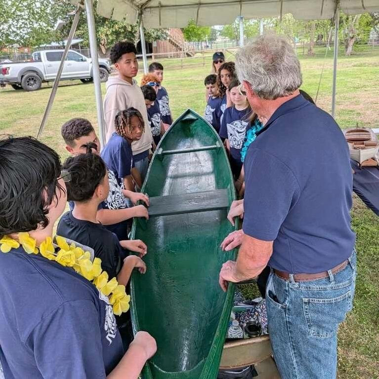 Group of children and an adult gathered around a green canoe under a canopy outdoors learning about traditional Louisiana boat building with Robert Theriot.