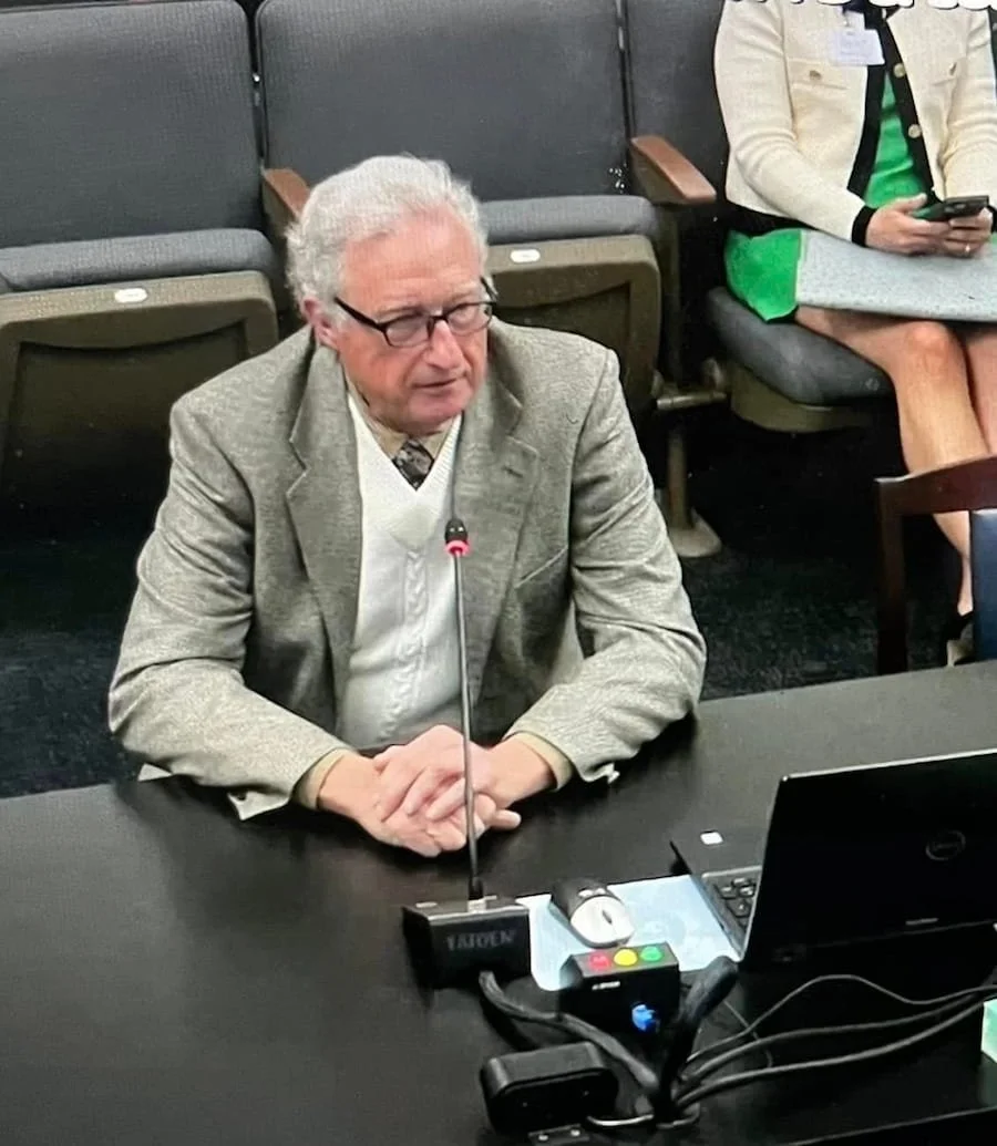 An older man with gray hair and glasses, wearing a gray blazer, is speaking into a microphone during a formal meeting or conference. He is seated at a table with a laptop, a mouse, and some electronic devices. There are other attendees in the backgro