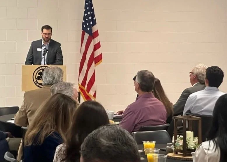 A man in a suit and glasses giving a speech at a podium with a logo, behind an American flag, in a room with seated audience members, some with drinks on the table.