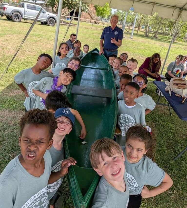 Children and adults gathered around a green wooden canoe under a canopy outdoors at a daytime event learning about traditional Louisiana boat building with Robert Theriot.