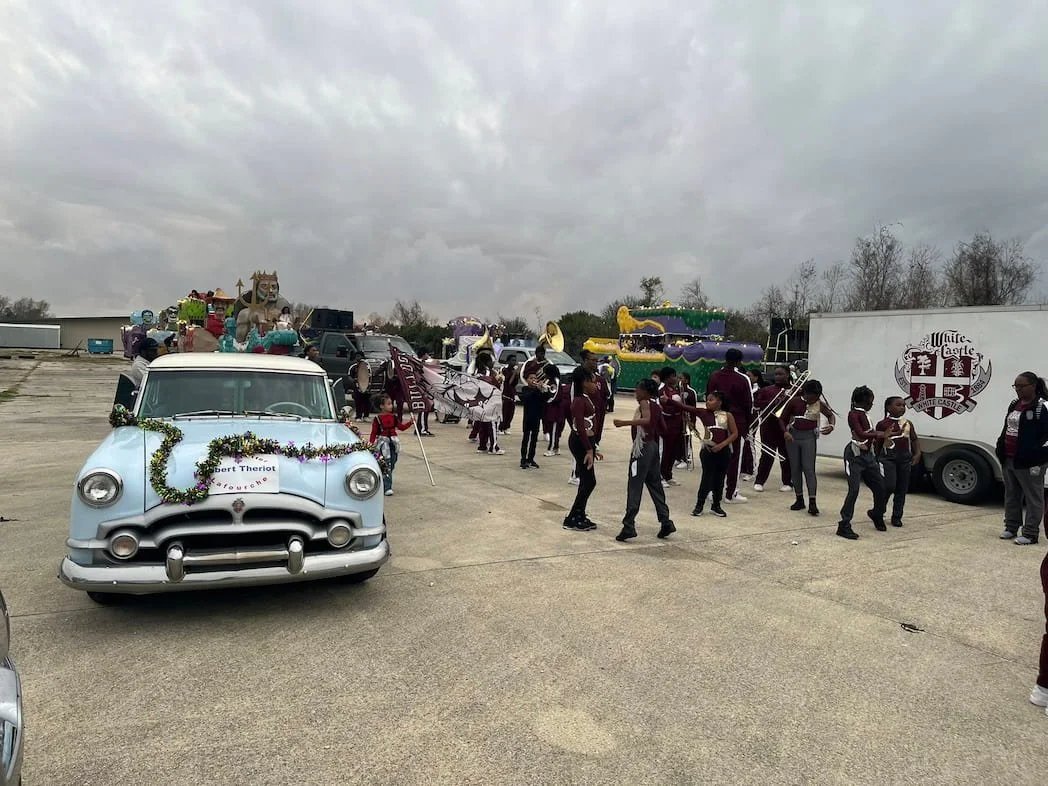 A parade scene on a cloudy day with a group of children and adults, some in maroon uniforms, walking and playing instruments near decorated floats like a vintage car with floral decorations and a castle-themed float in the background.