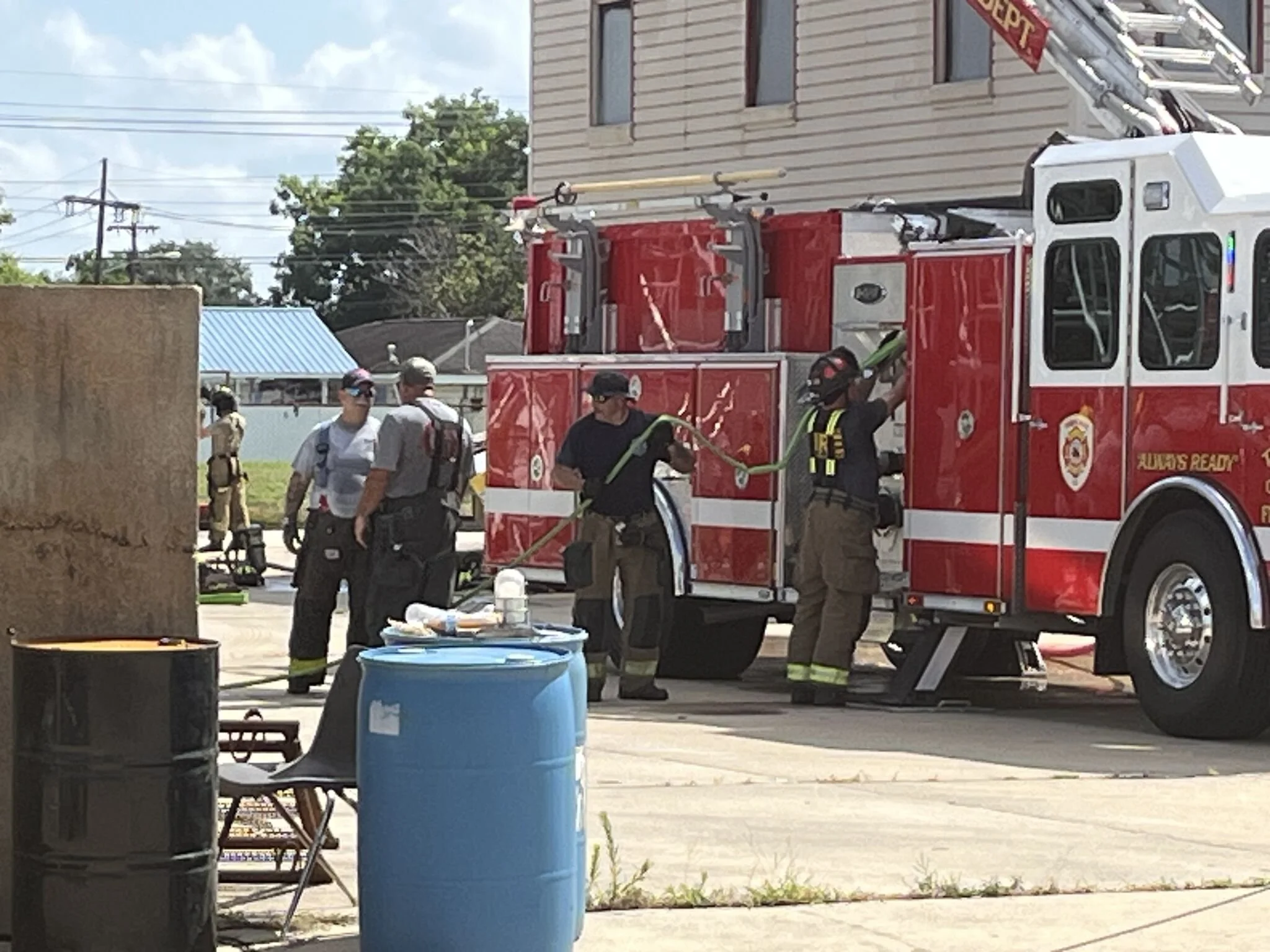 Firefighters and emergency responders gather around a red fire truck on a street, with emergency equipment and blue barrels nearby. There are houses, trees, and utility poles in the background.