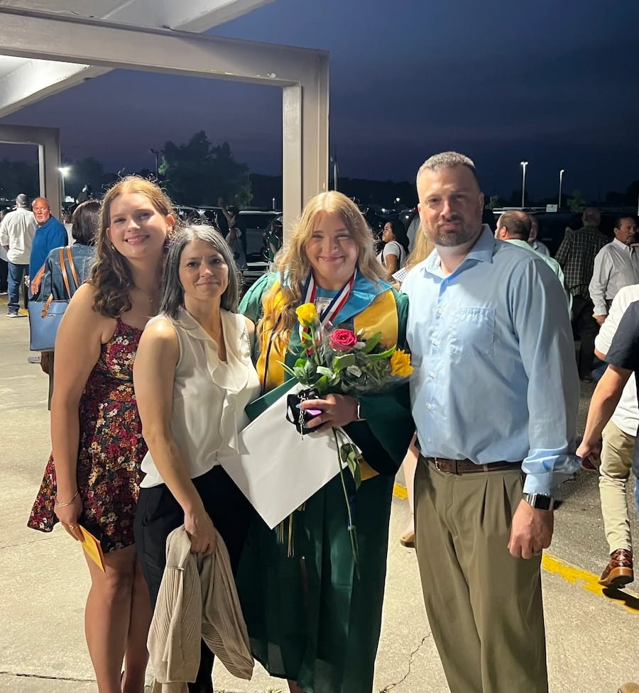 A group of four people, including a woman in a graduation gown holding flowers and smiling, standing at an outdoor evening event with other people around.
