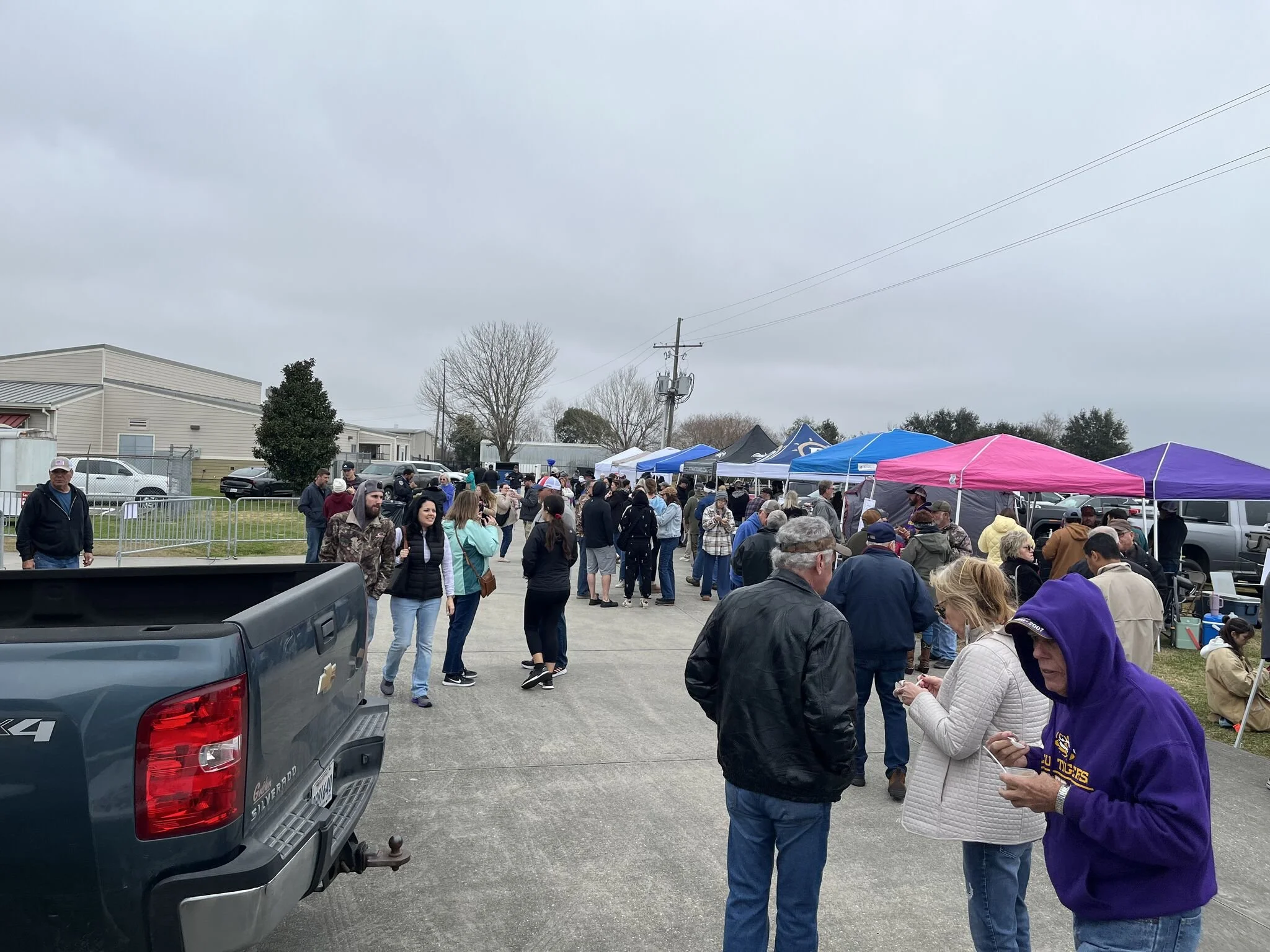 A crowd of people gathered outdoors under overcast skies, standing near tents and a parking lot with vehicles, with power lines overhead.