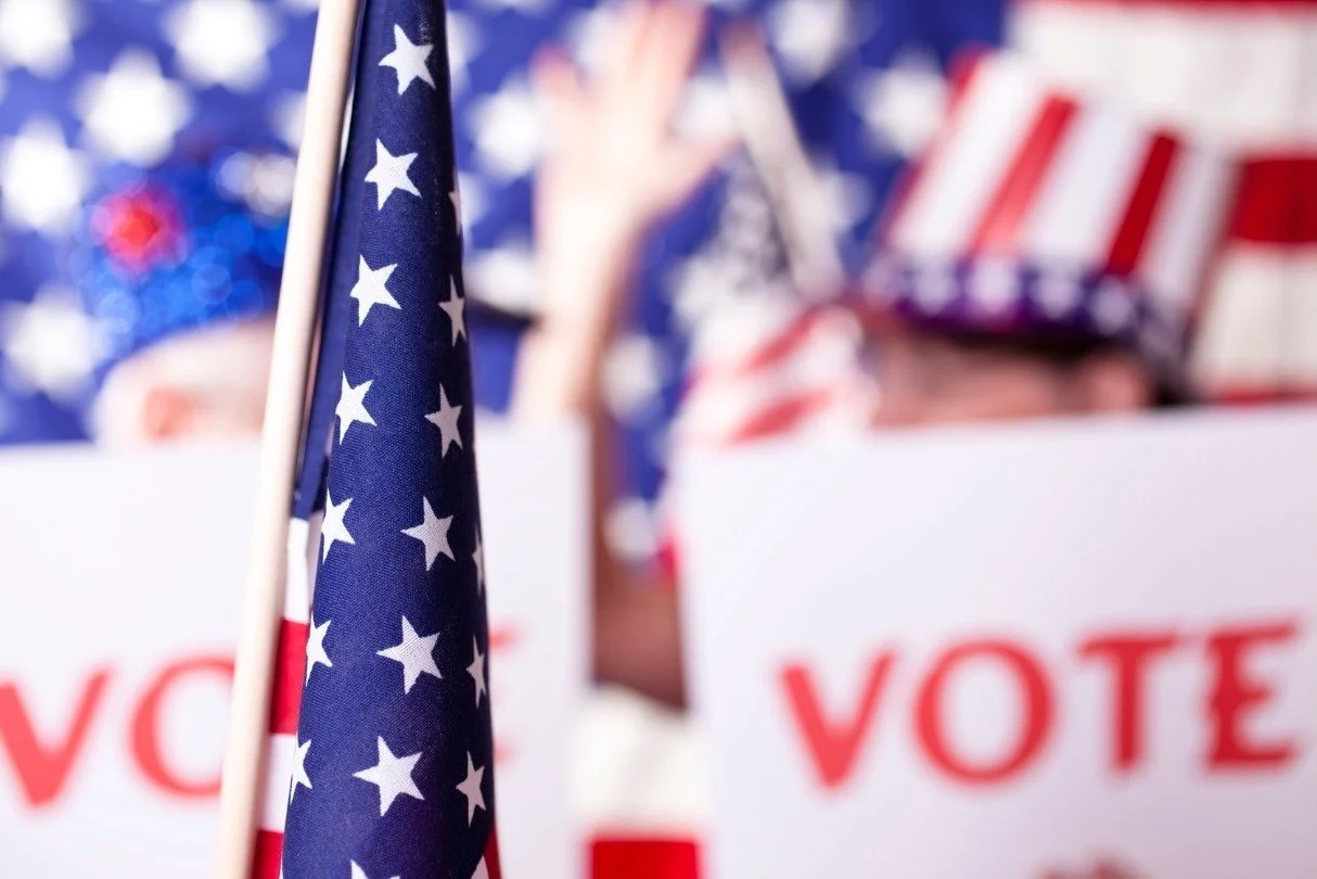 Close-up of an American flag at a voting event with a person in the background wearing patriotic attire and holding a sign that says 'VOTE'.