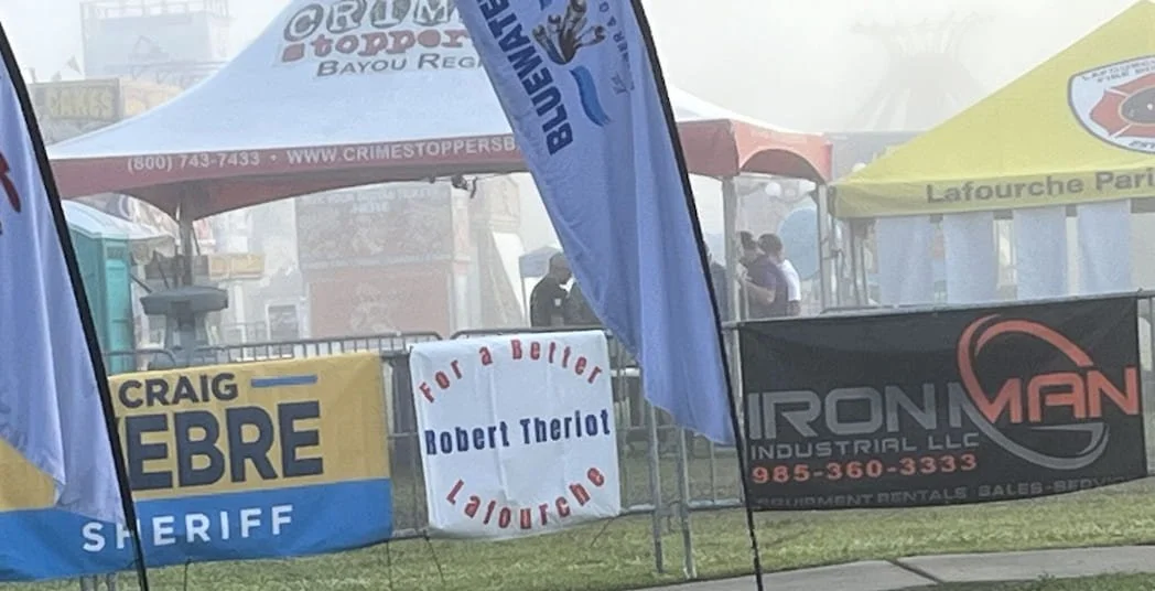 At an outdoor event with booths and tents, including banners for Crime Stoppers, Lefourche Parish, Craig Lebre Sheriff, Robert Theriot for a better Lafourche, and Ironman Industrial LLC, with people visible in the background.