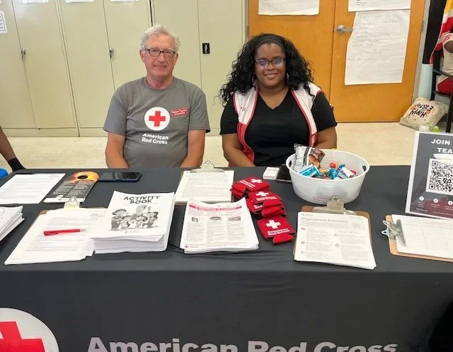 Robert Theriot sitting behind a table with American Red Cross materials, one man with gray hair and glasses wearing a Red Cross t-shirt, and a woman with curly black hair in black and red clothing, in an indoor setting.