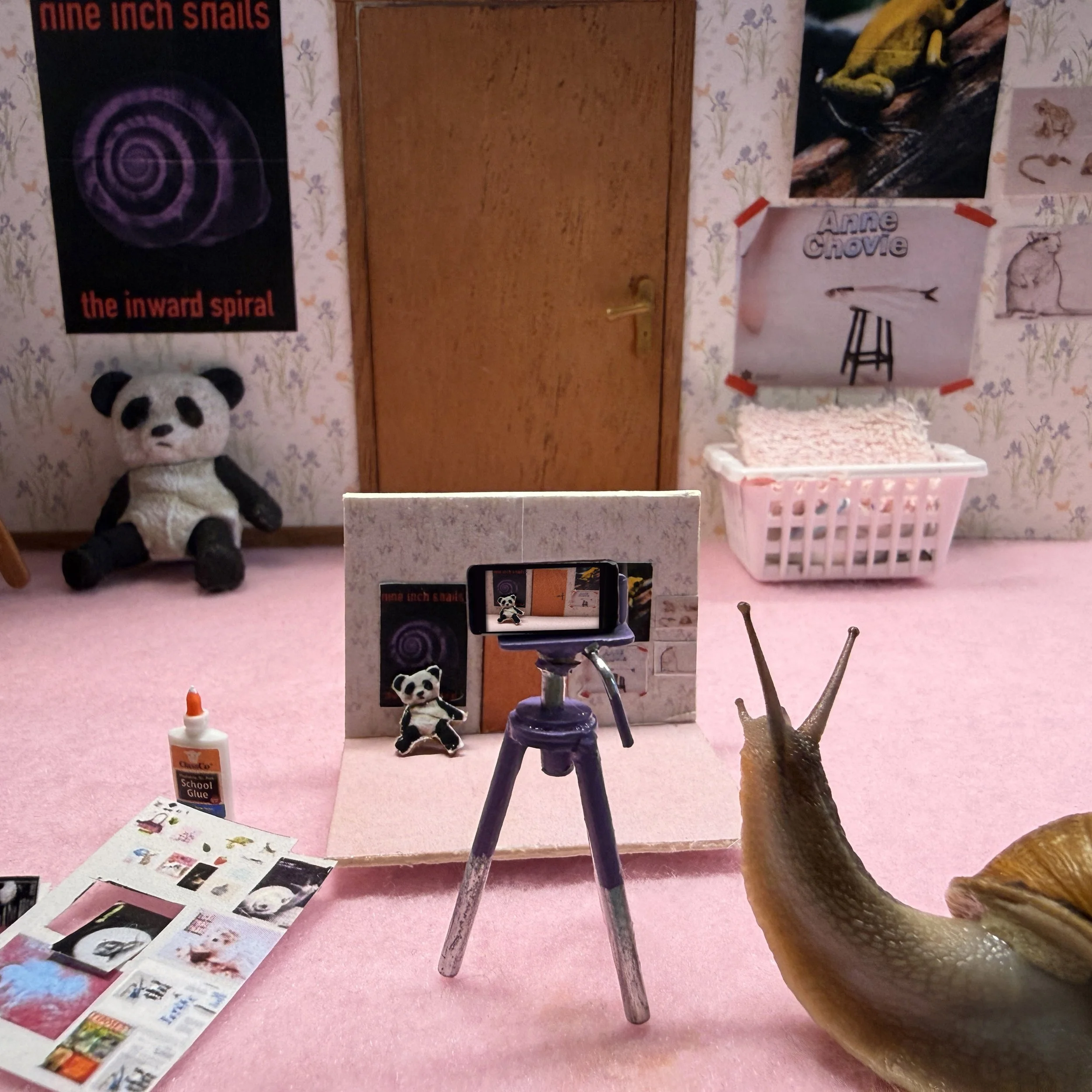 Room decorated with animal-themed posters and toys, including a panda plush. There is a variety of paper crafts on the pink carpeted floor. Velveeta the snail is documenting their miniature project.