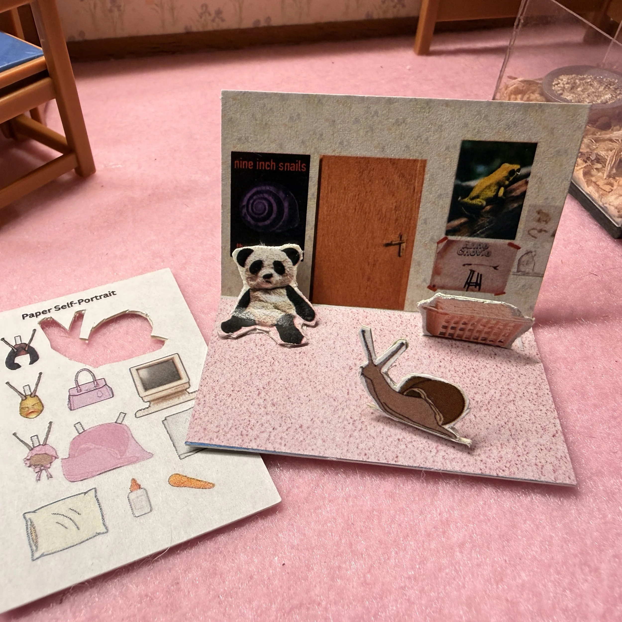 A pink carpeted floor with a cardboard miniature of the larger bedroom.