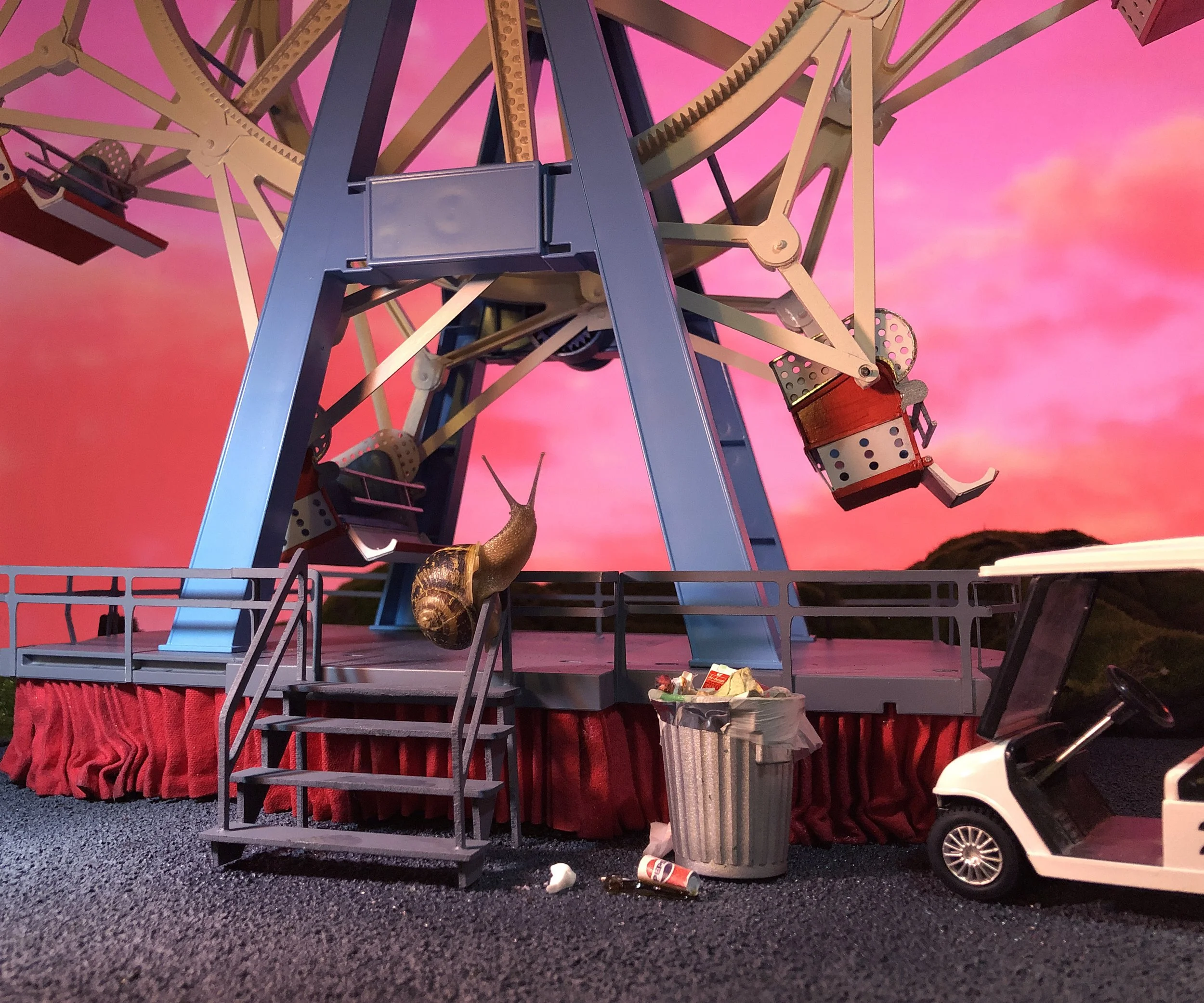 Close-up of a Ferris wheel at dusk against a pink sky with a small toy car in the foreground and litter on the ground. A snail is seen in the foreground of the carnival.