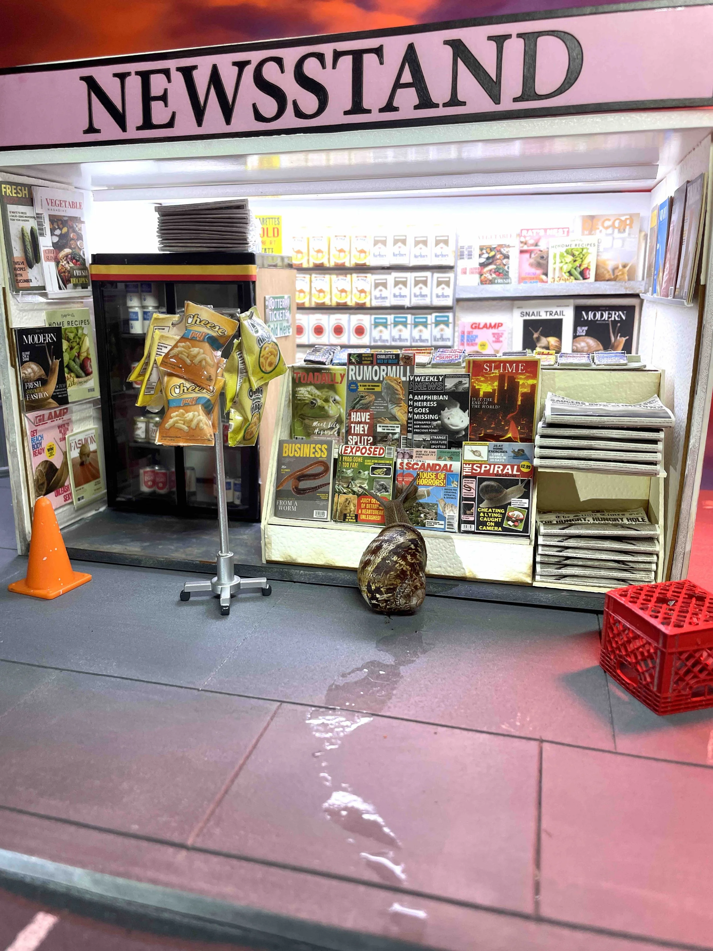 Close-up of a newsstand with magazines and snack bags in front, orange cone and red crate on the ground, with wet floor and water spill in front of the stand. A snail is at a newsstand at night.