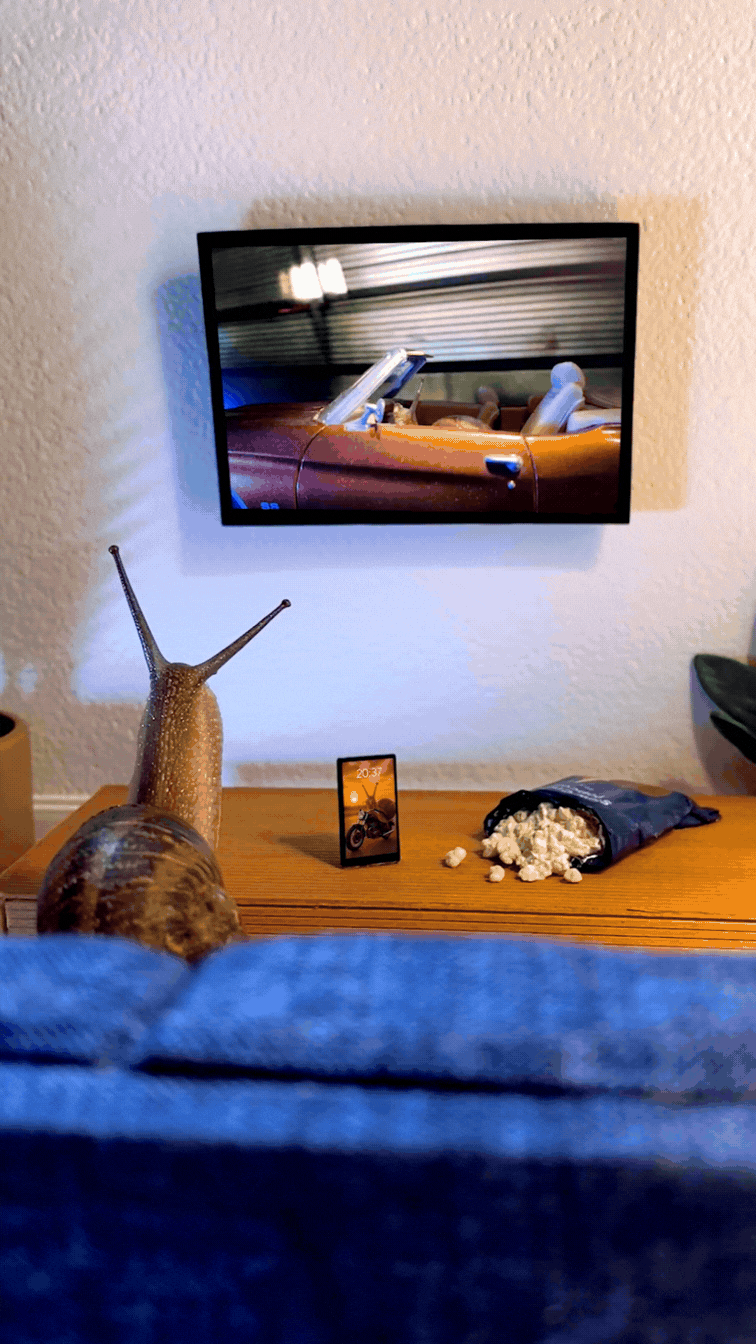 Living room with a snail watching an action movie with a big bag of popcorn, and a wall-mounted television showing a red sports car in a high chase getaway scene.