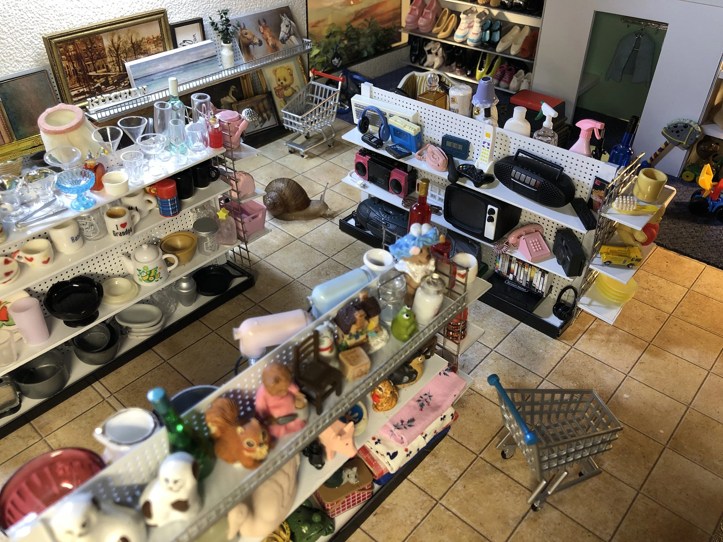 A miniature handmade thrift store. The shelves are filled with various glassware, ceramic mugs, and decorative items. In the background, there are decorative objects and art. In the back, a snail (shopping) on the tiled floor.
