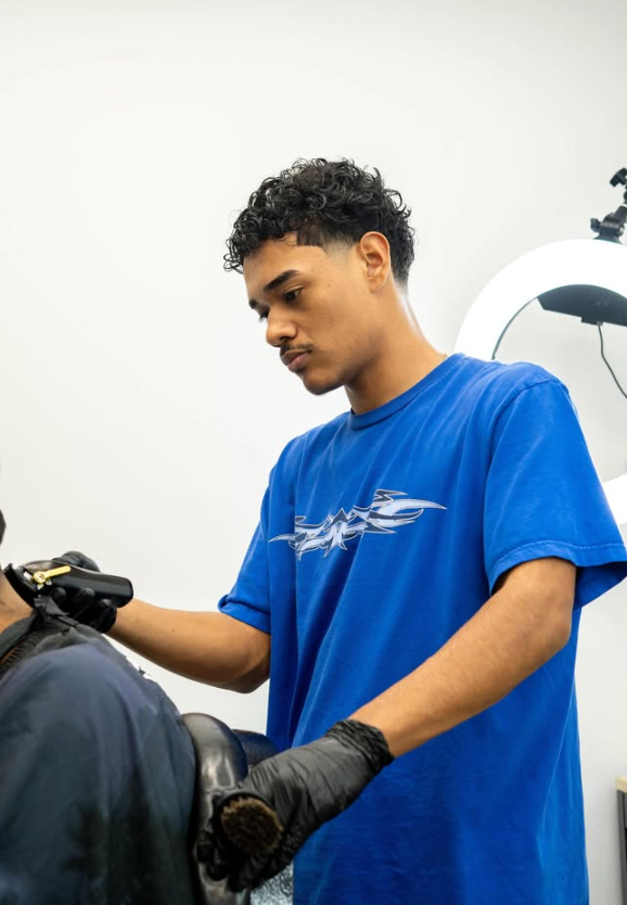 A young man with curly hair wearing a blue t-shirt and black gloves working on a patient in a clinical setting, possibly a medical or dental clinic.