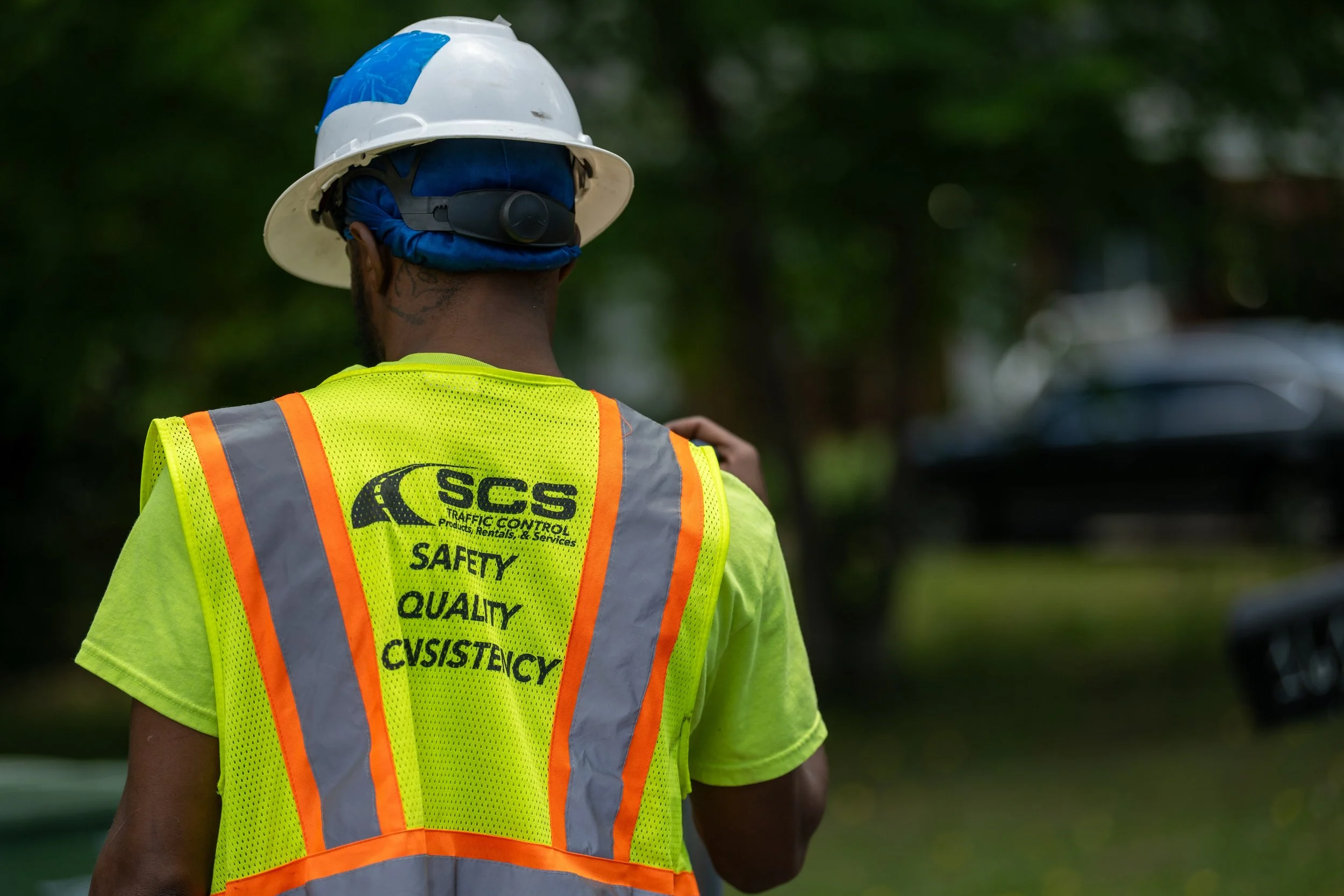 Back of a construction worker wearing a white hard hat, blue head covering, and a yellow safety vest with orange and gray reflective stripes. The vest has text related to traffic control, safety, quality, and consistency.