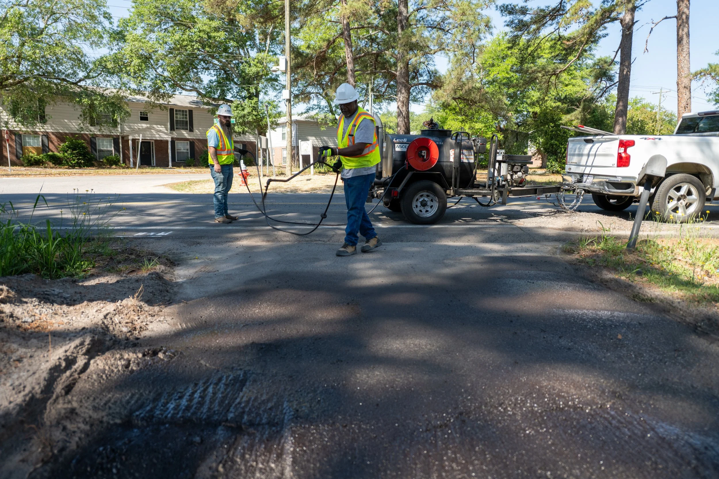 Workers in high visibility vests and helmets repair a road using heavy equipment, with a residential neighborhood and trees in the background.