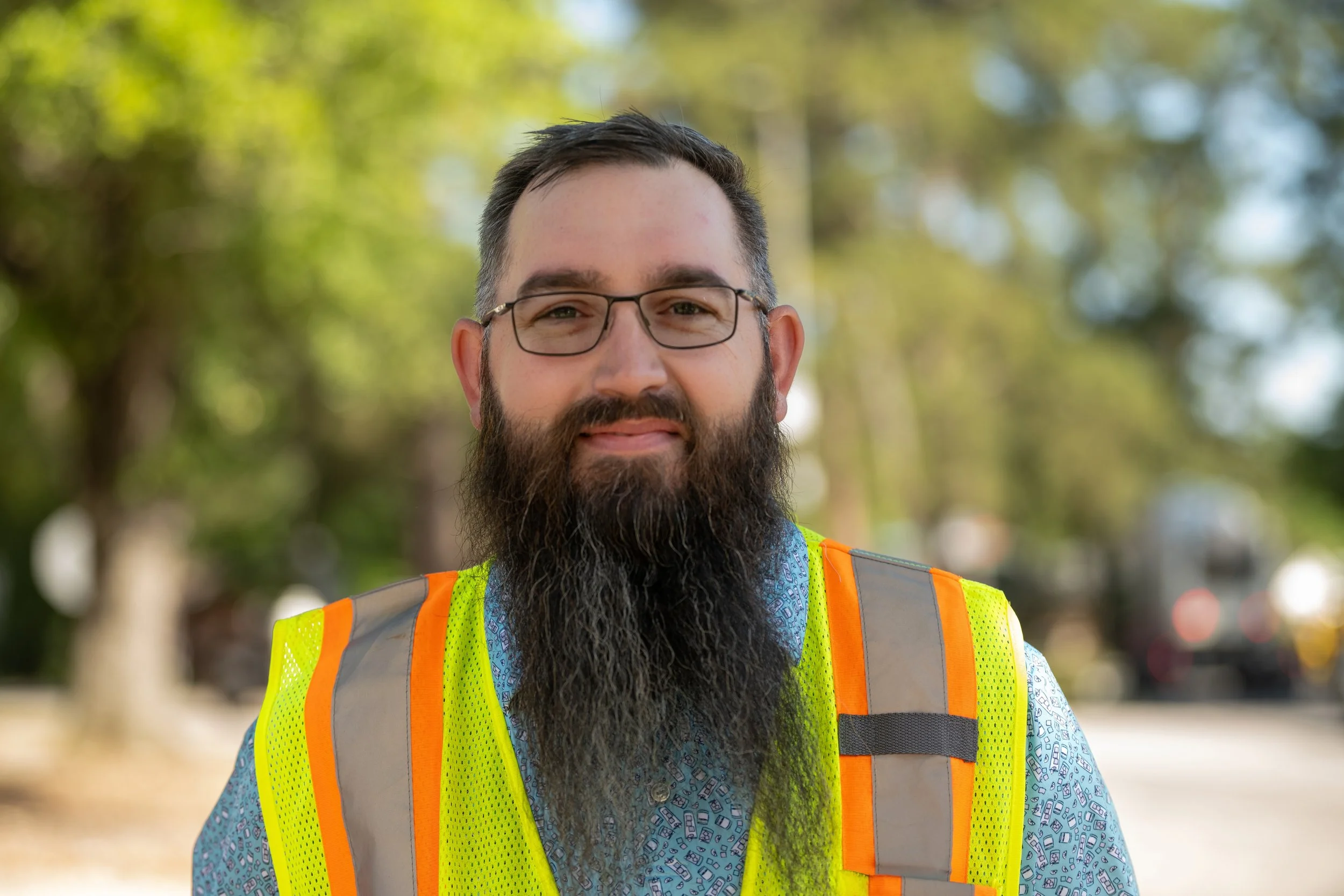 Man with glasses and a beard wearing a high-visibility safety vest outdoors, with a blurred background of trees and sunlight.