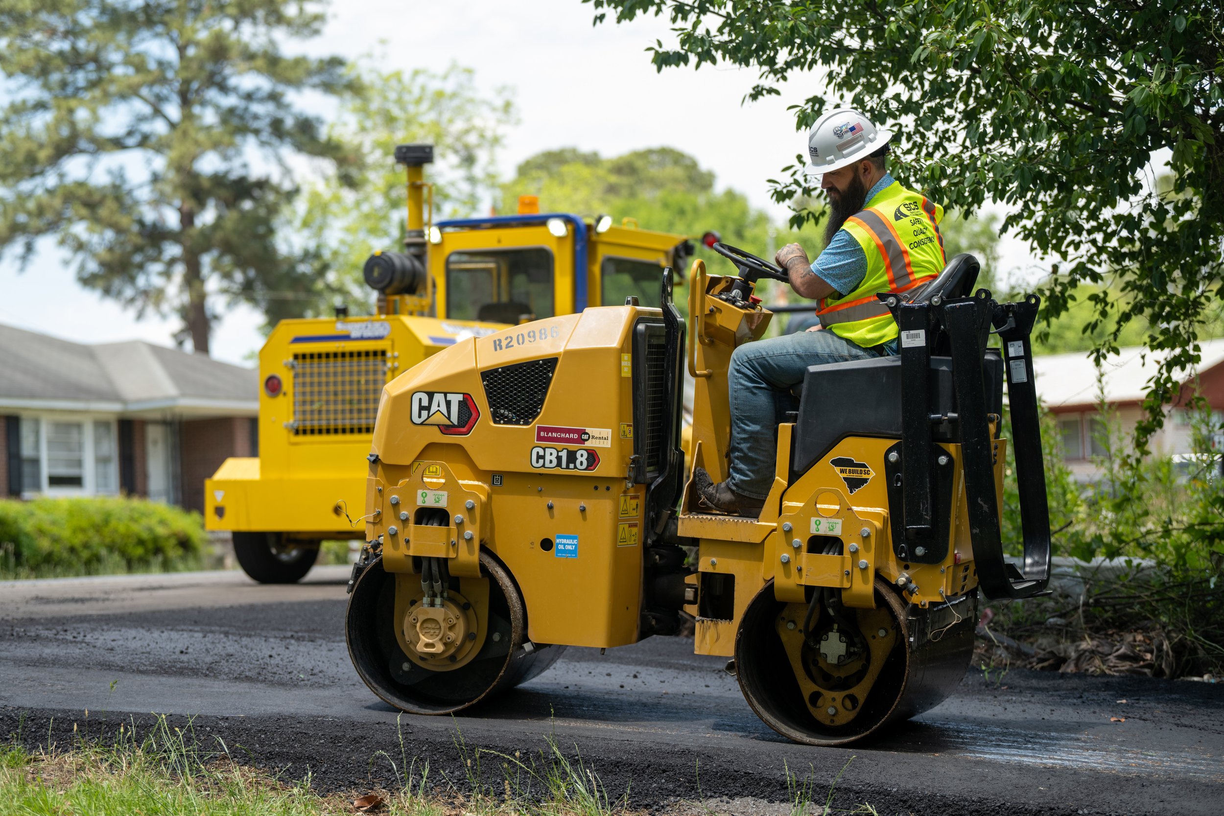 A construction worker operating a yellow CAT asphalt roller on a residential street with trees and houses in the background.